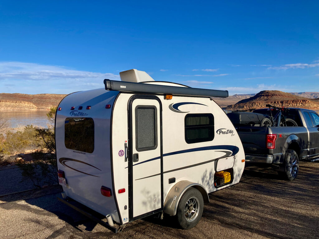 Small white trailer beyond charcoal grey truck in campsite above distant lake.