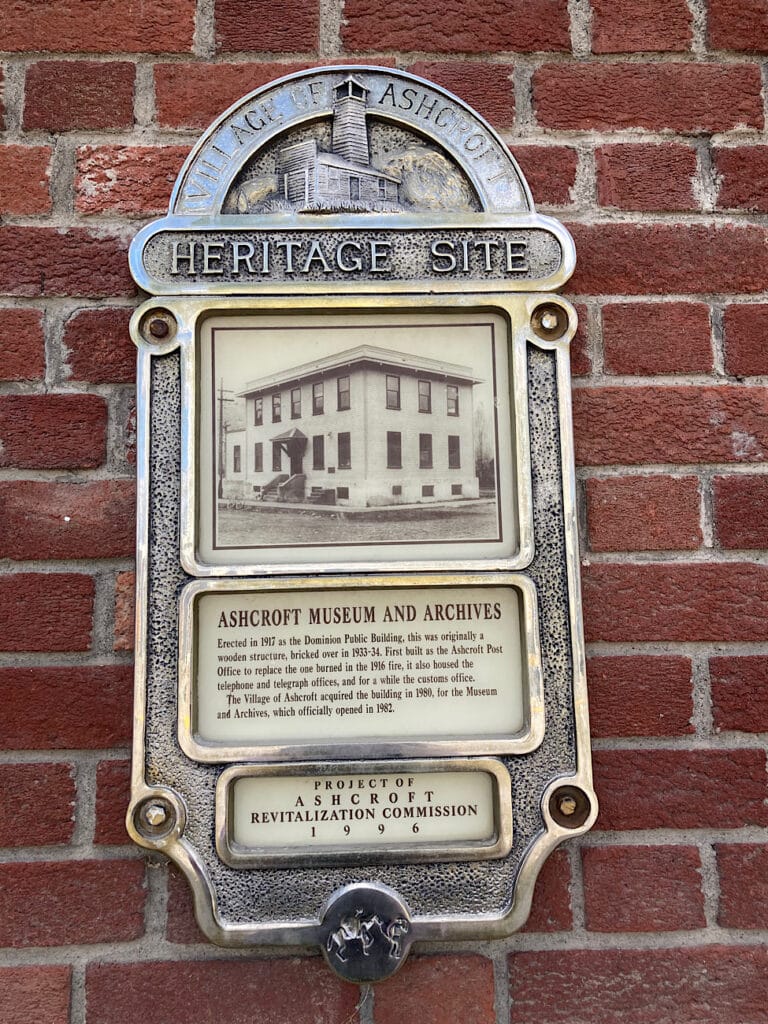 Metal plaque on brick building reading: Ashcroft heritage site (and details of building history).