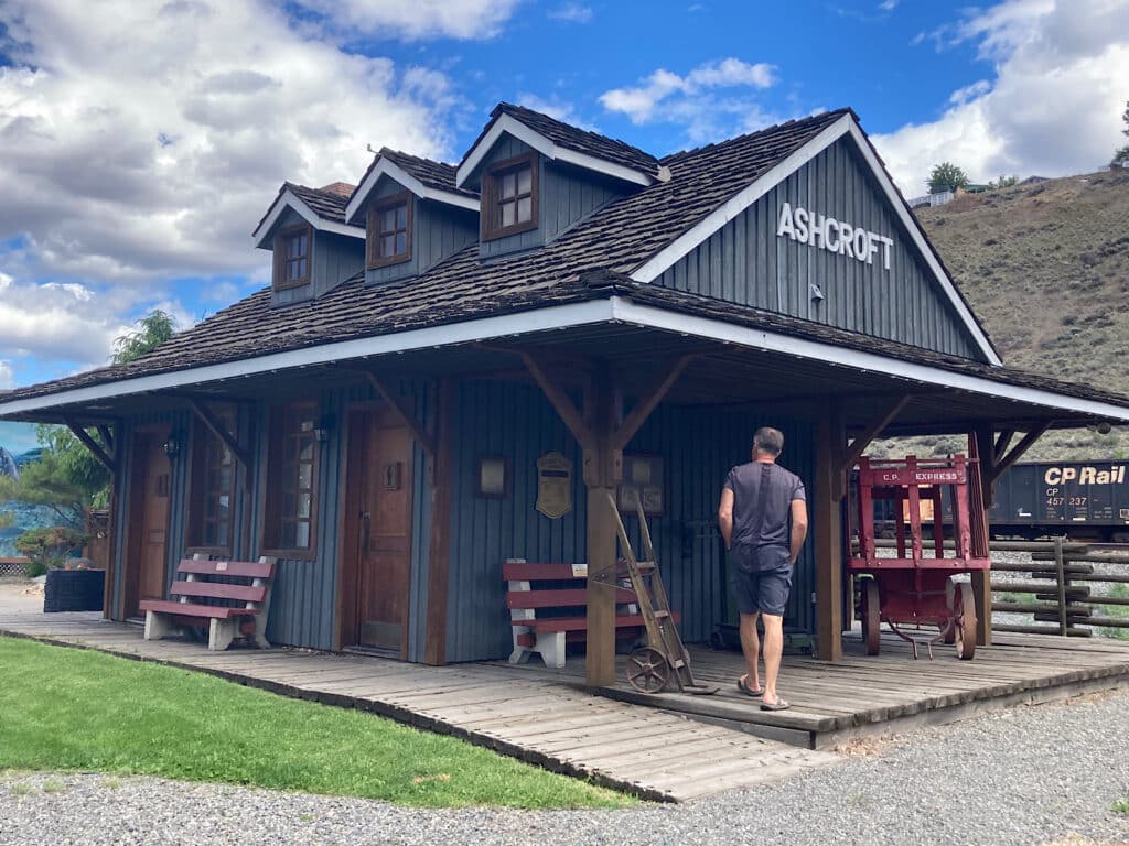 Man walking towards historic equipment on porch of grey building next to a rail line.
