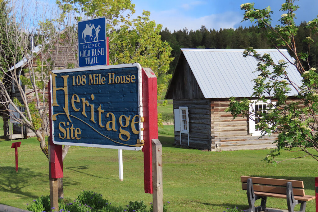 Entrance sign reading: 108 Mile Heritage Site, with wooden building behind.