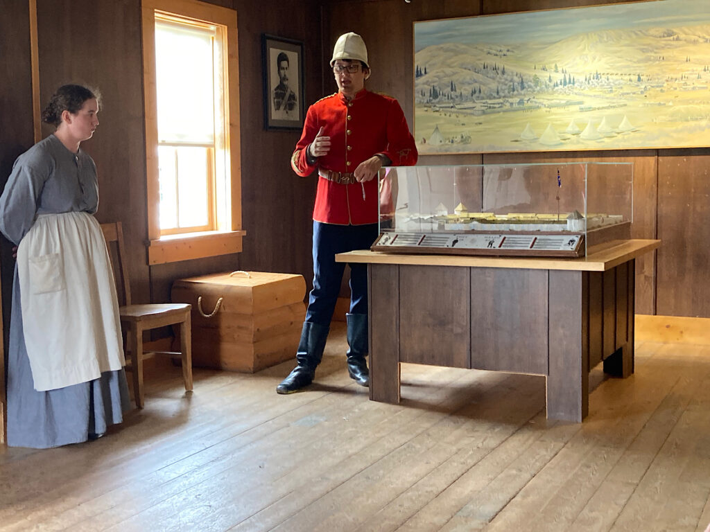 Man in red jacket and dark pants standing by diorama while woman in grey dress and white apron stands to one side.