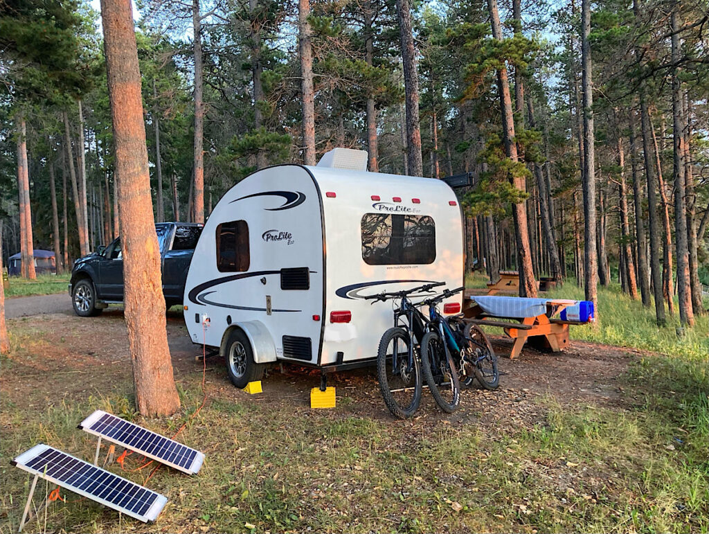 Small trailer set up in wooded campground with 2 solar panels set up nearby.