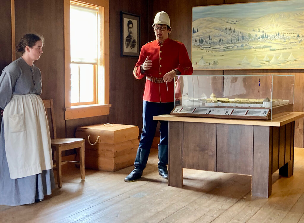Man in red tunic and dark trousers standing by a model while woman in long grey dress and white apron stands watching.