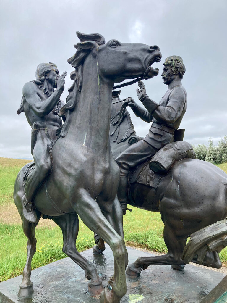 Statue of Indigenous man and man in uniform passing each other on horseback.