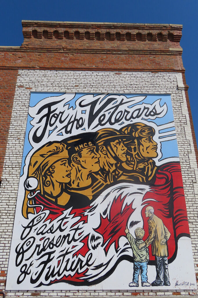 Painted mural of old man and young boy looking on large Canadian flag with heads of veterans above.