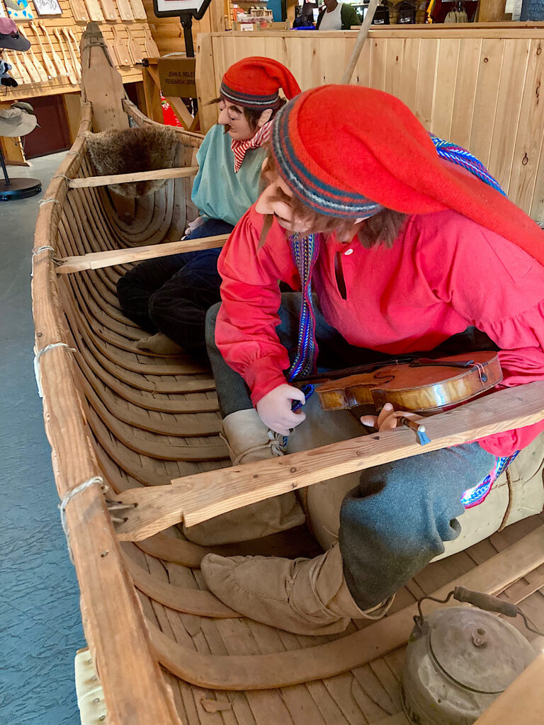 Two mannequins dressed in red clothing in a canoe with musical instruments.