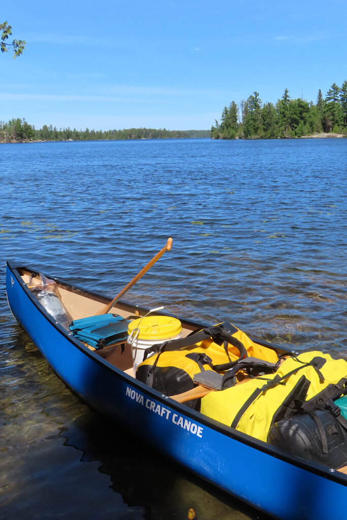 Blue canoe sitting on lakeshore loaded with yellow bags and other gear.