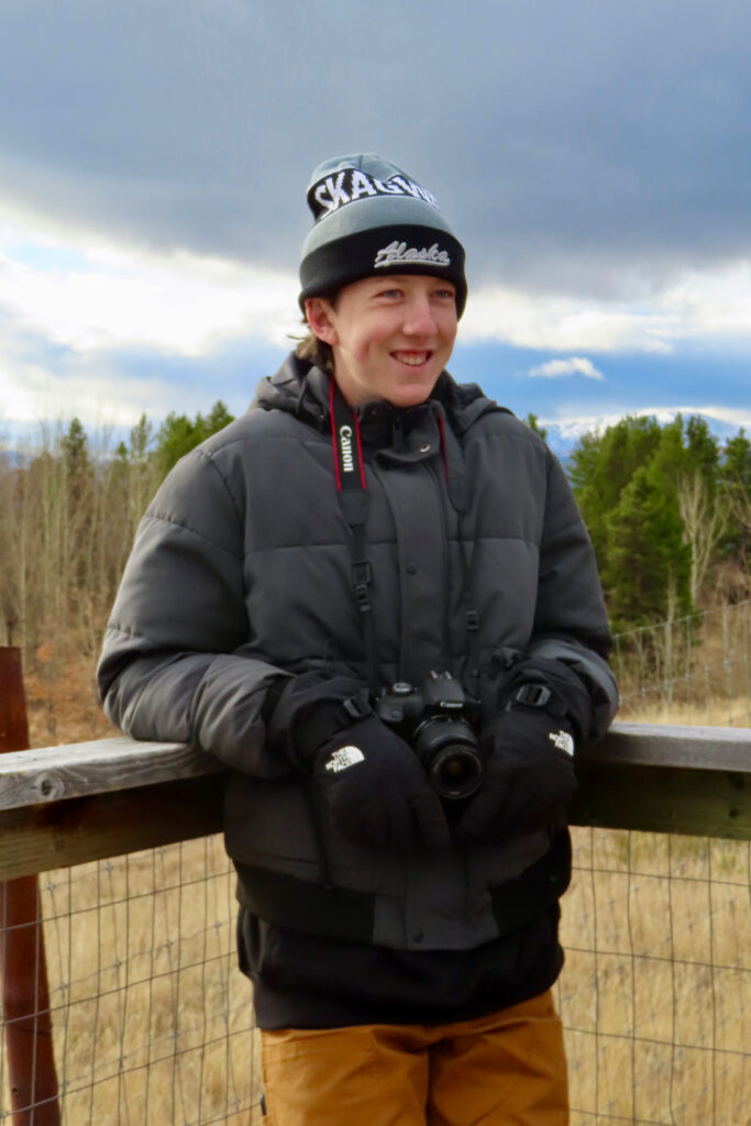 Young man in jacket, hat and mitts with camera leaning against a wooden fence.