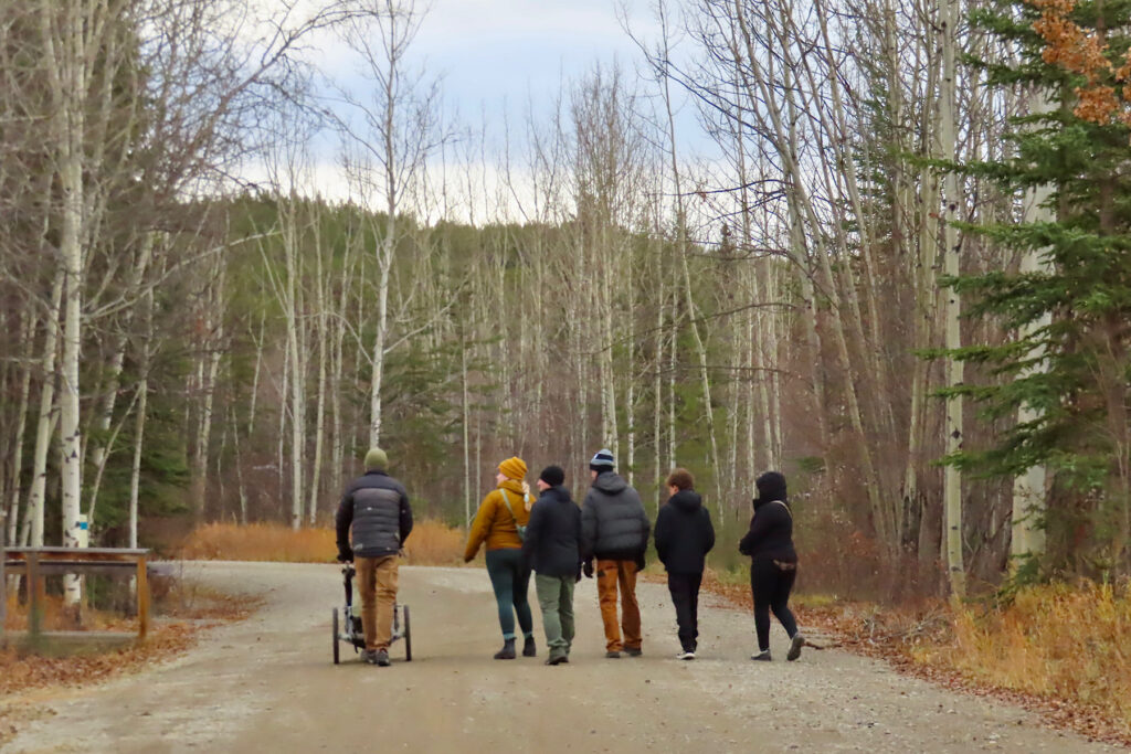 Group of 6 people plus a stroller walking down gravel road in a forest.