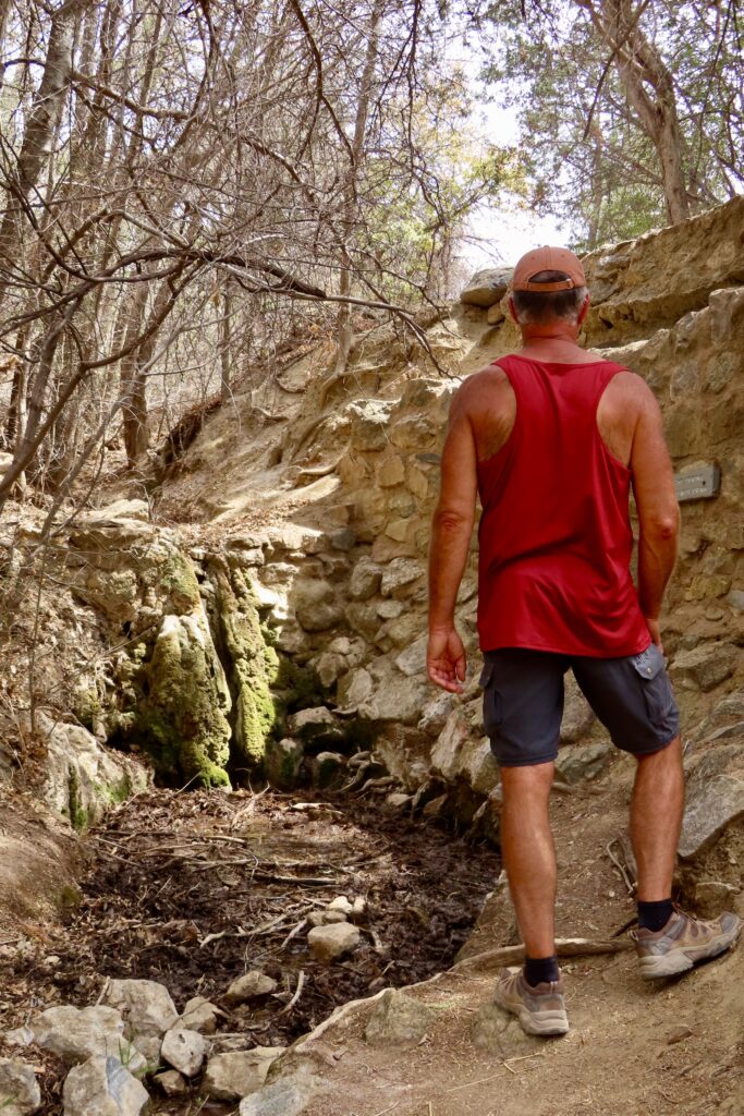 Man in red shirt and dark shorts standing looking at mossy spring.