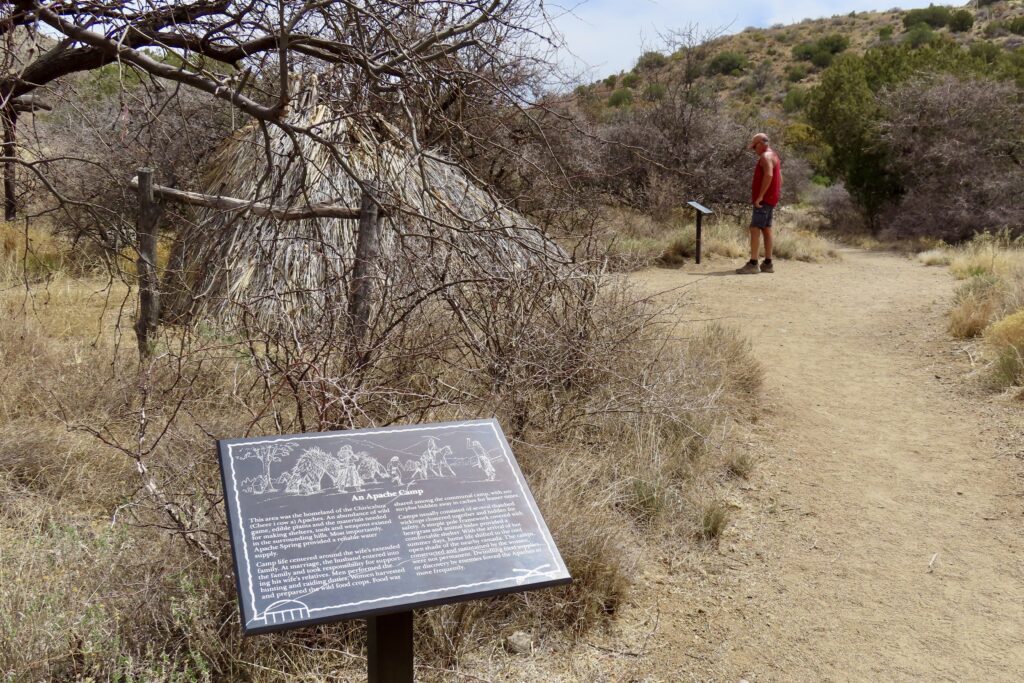 Interpetive sign for Apache Camp with man standing outside of replica wigwam-style structure.