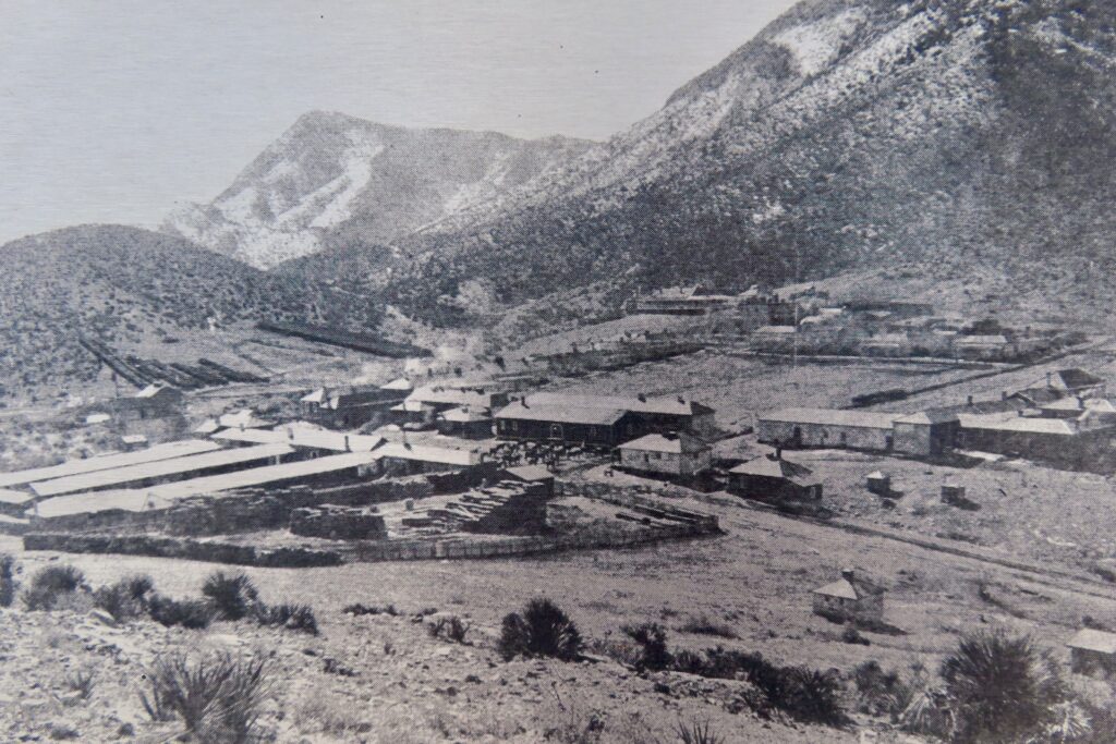 Black and white photo of old fort on flat ground in front of mountains.