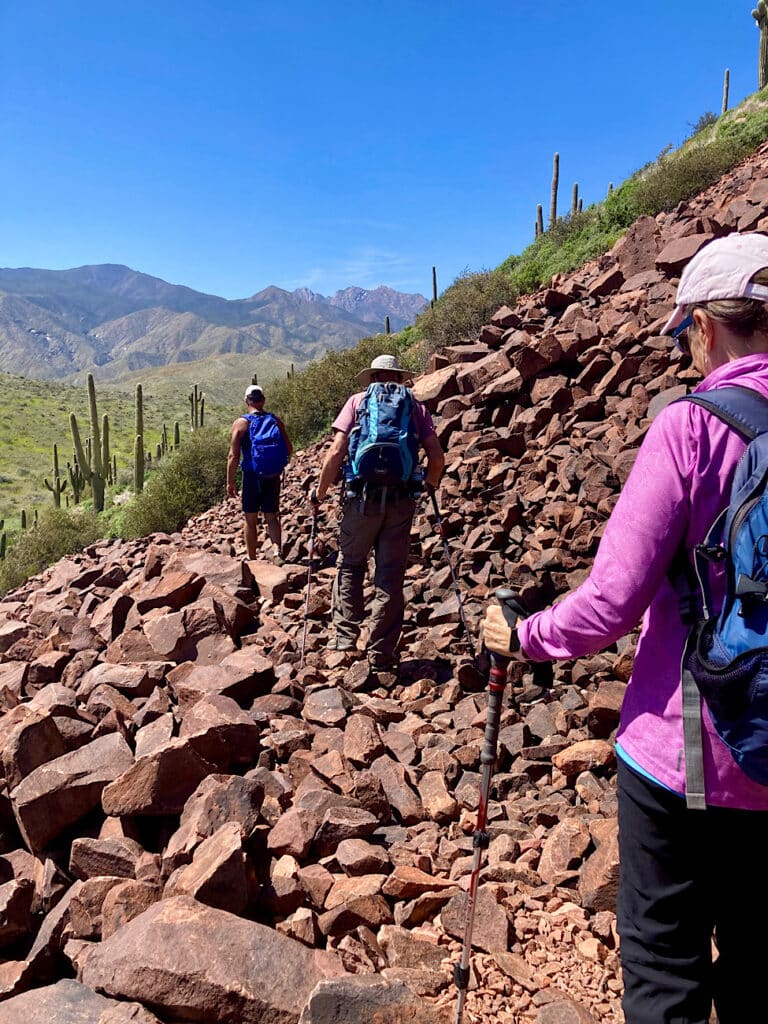 Two men and a woman in pink jacket walking across rubbly rock pile towards distant mountains.