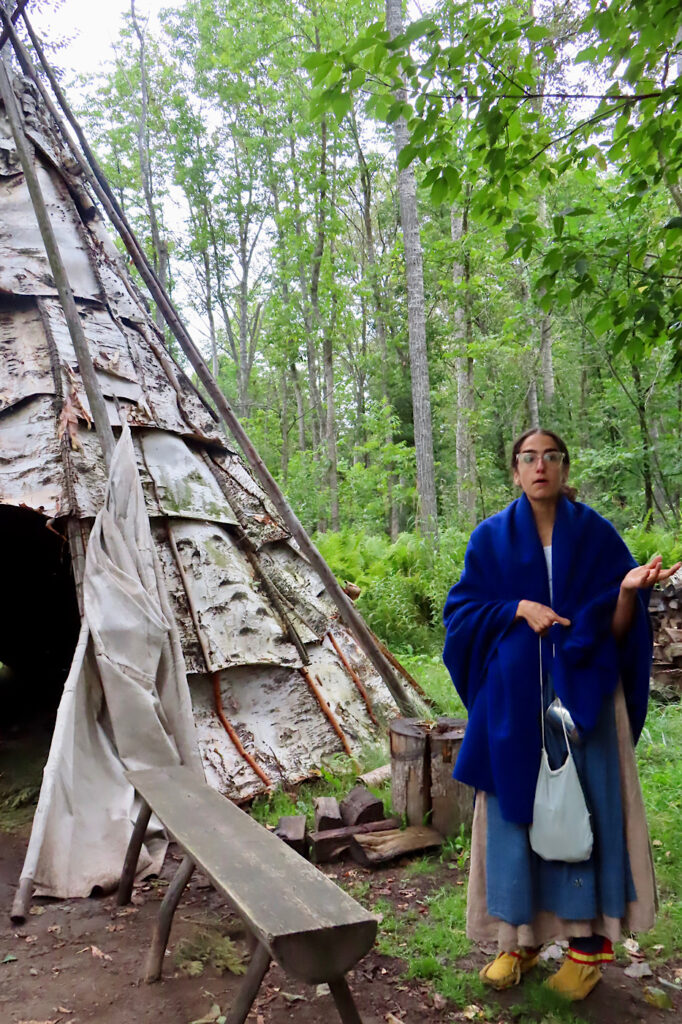 Woman in blue shawl and dress wearing moccasins standing outside wooden structure.