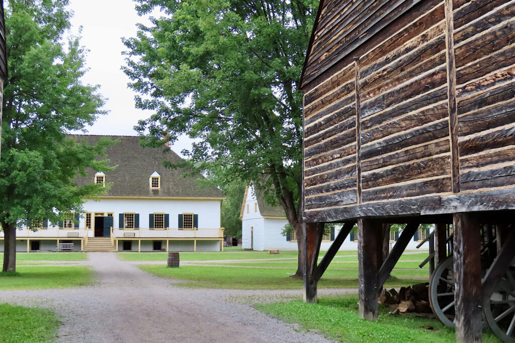 Raised wooden building on right and whitewashed building ahead of green grassy area.