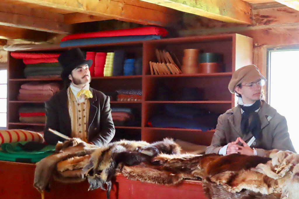 Men dressed in period costumes standing behind counter with shelves filled with fabric and sewing materials behind them.