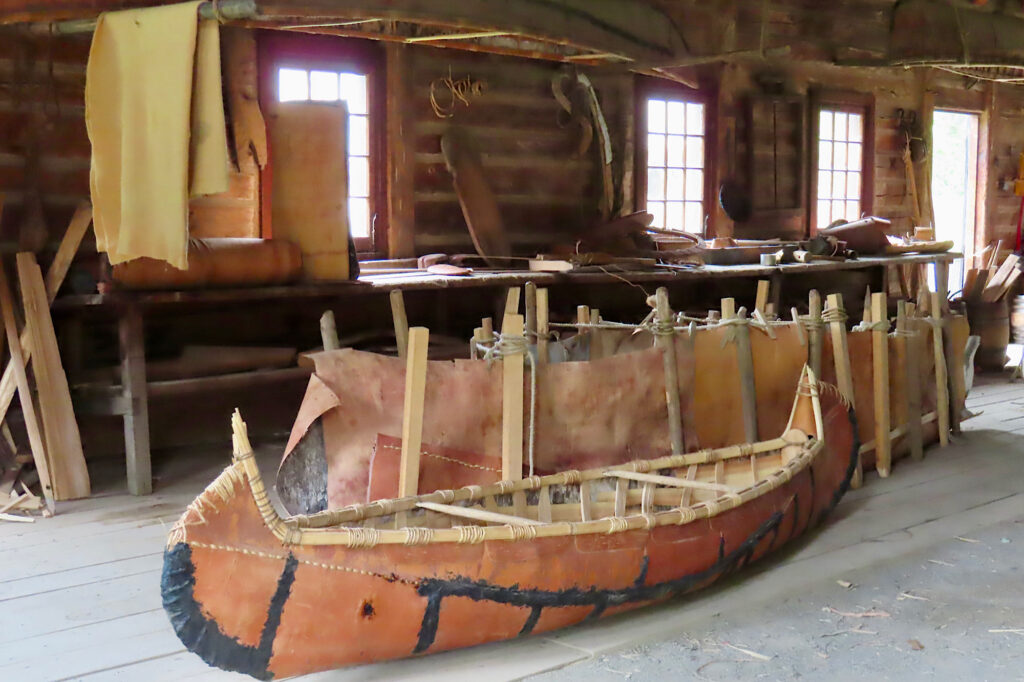 Birchbark canoe in shed with another under construction behind.