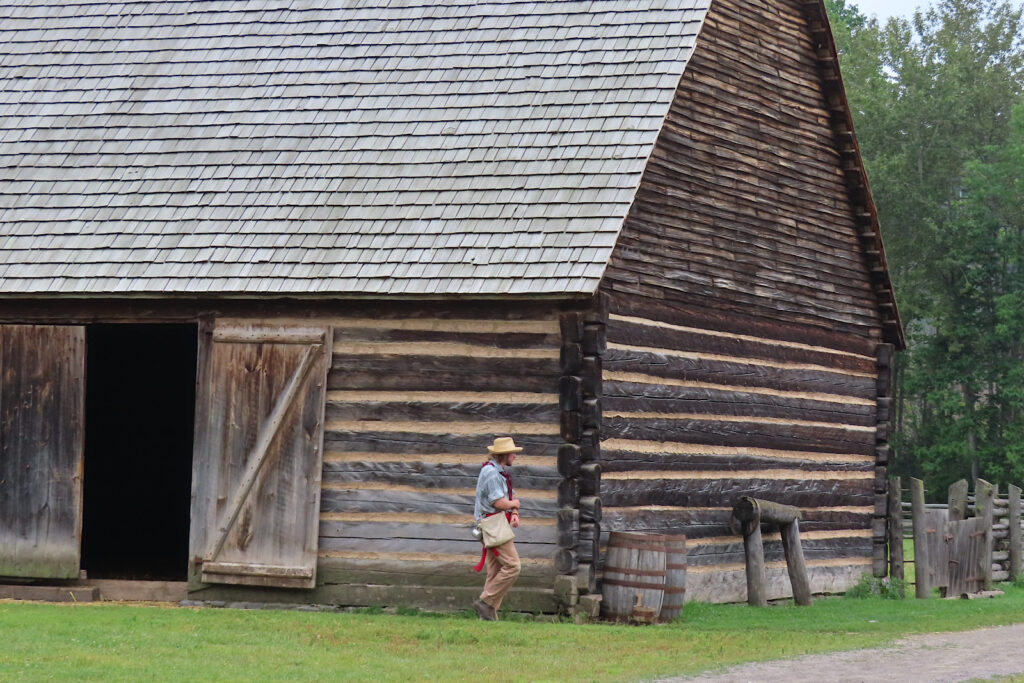 Man in period costume walking past large wooden barn.