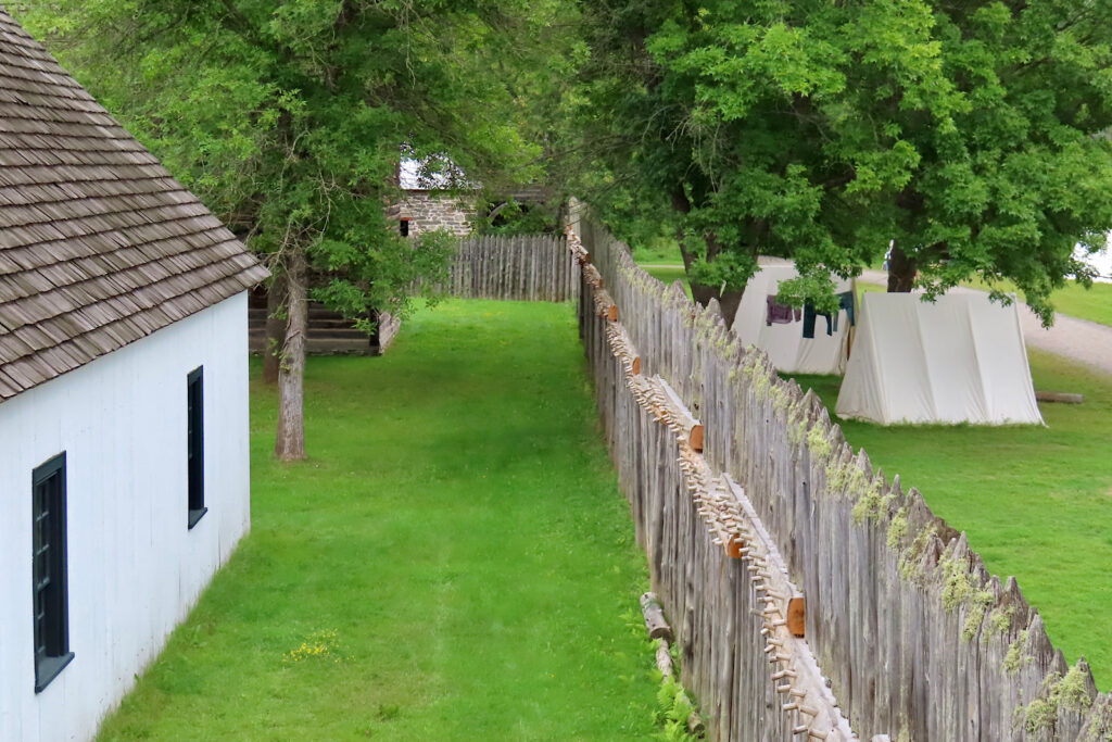 Wooden fort wall with whitewashed building on one side and tents on the other.