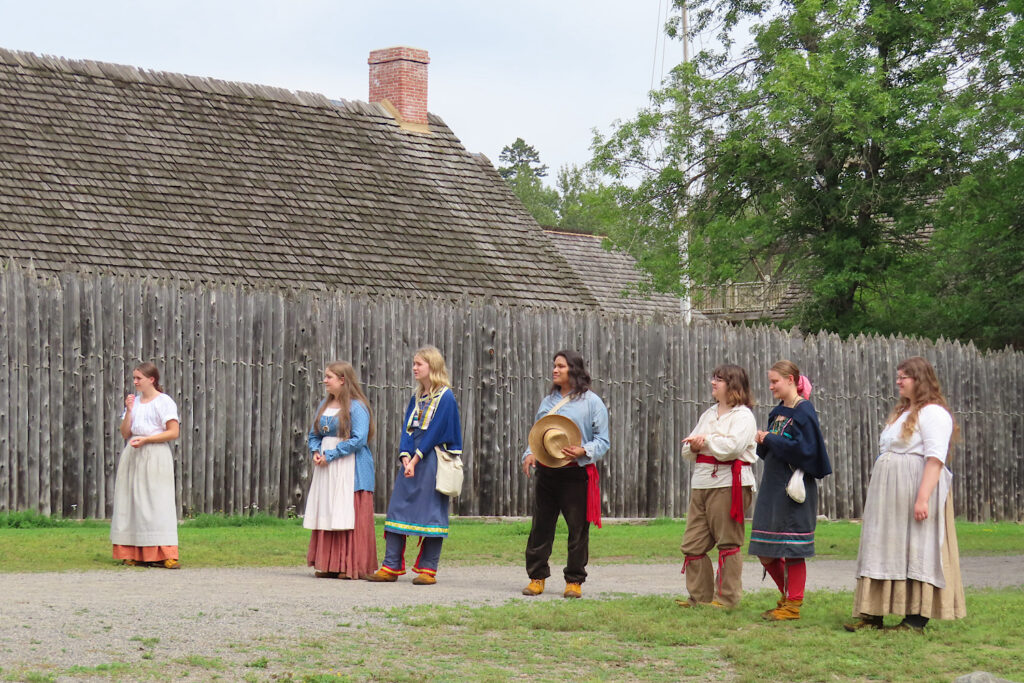 Group of people in historic costumes outside wooden fort wall.