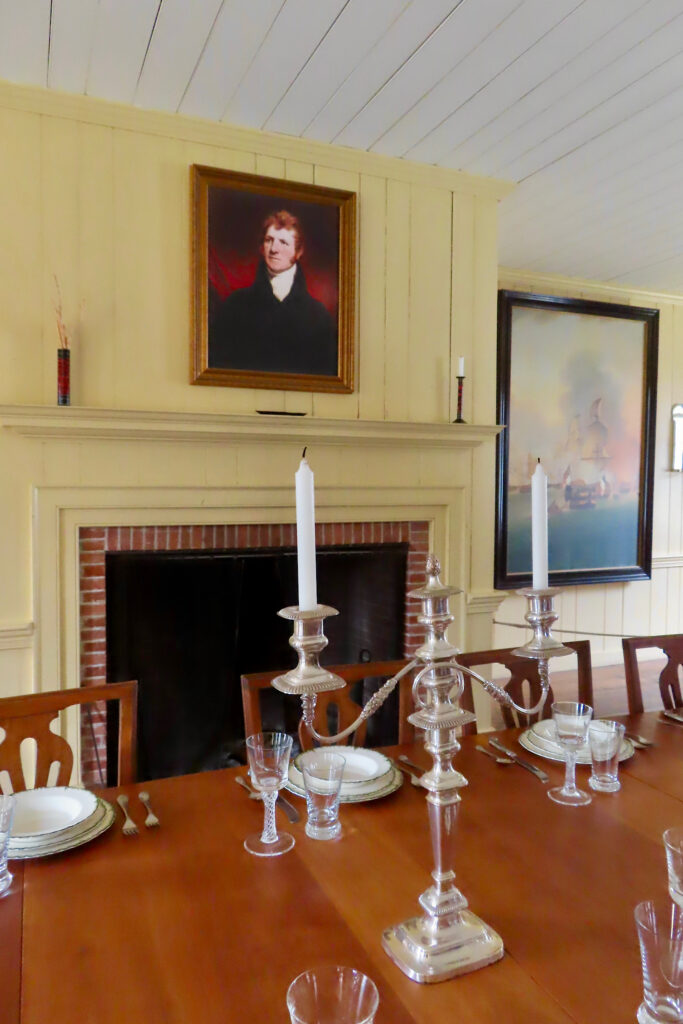 Wooden dining table with candlesitcks, wine glasses and china set in front of fireplace topped with portrait of a man.