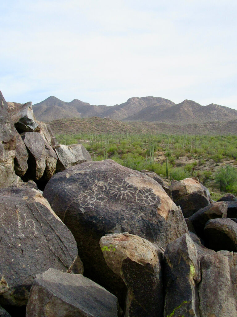 Piles of boulders with carvings in front of Saguaro cactus desert and distant mountains.