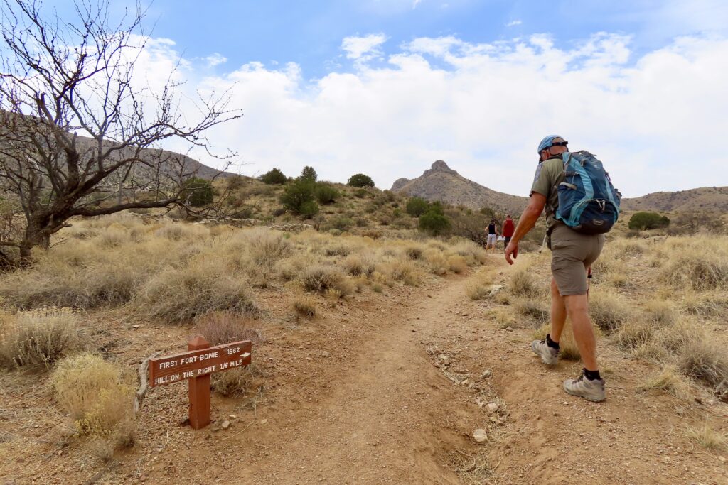 Male hiker in shorts carrying backpack walking trail towards two other hikers reading interpretive sign.