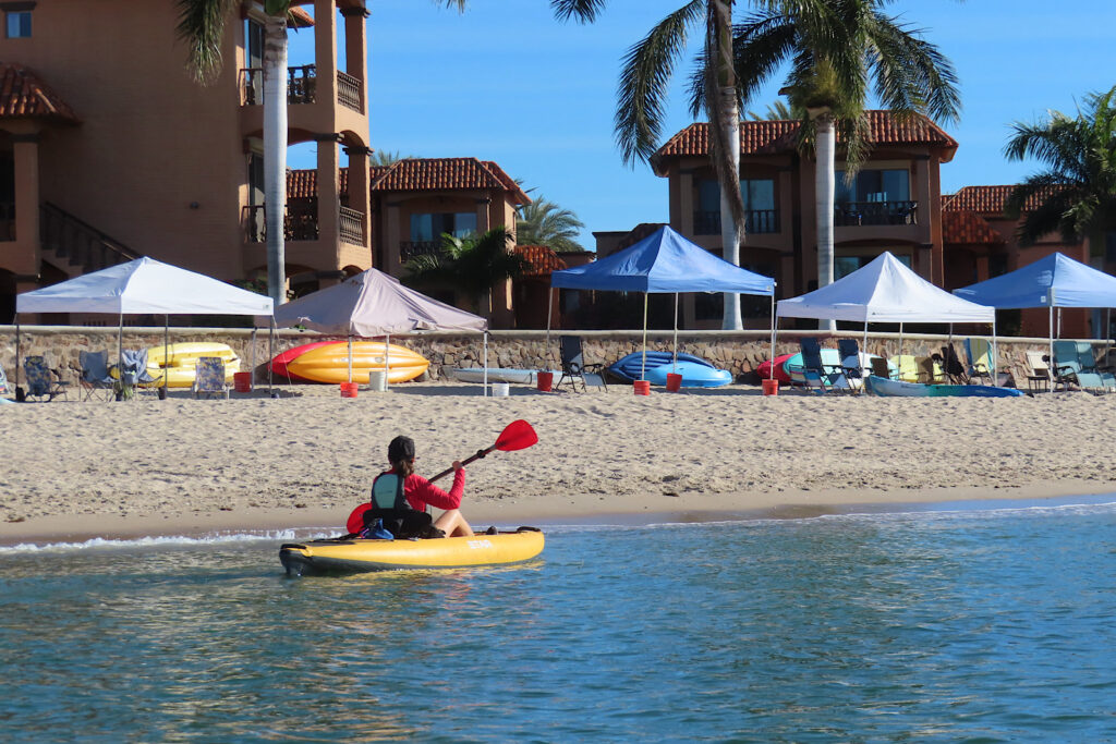 Woman in yellow kayak with red paddle passing sandy beach with sun shelters.