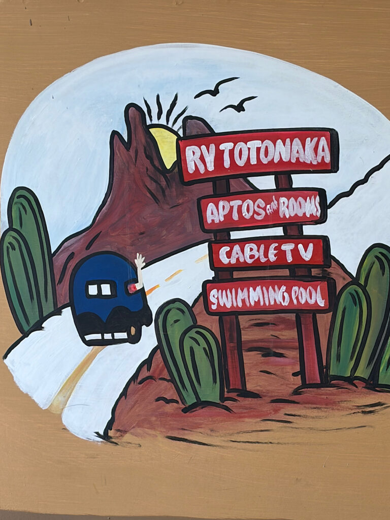 Painted sign of RV on road and sun setting behind mountain with words: RV Totonaka Apartos & Rooms, Cable TV, Swimming Pool.