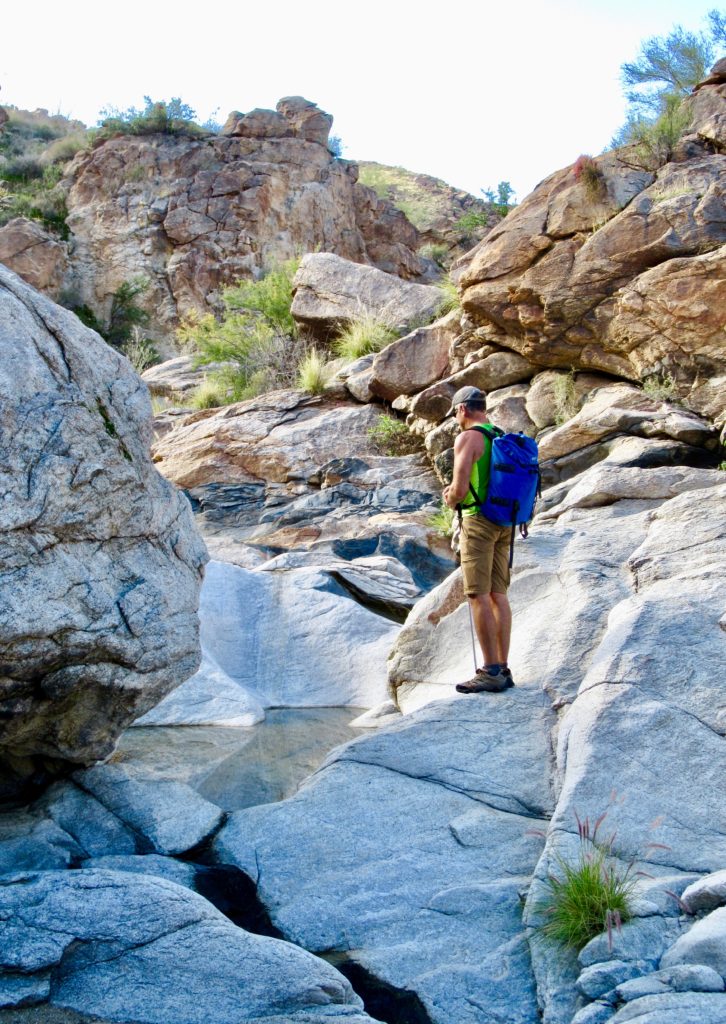 Man in green shirt, brown shorts and blue backpack stands in a canyon on smooth white rock above a pool of water.