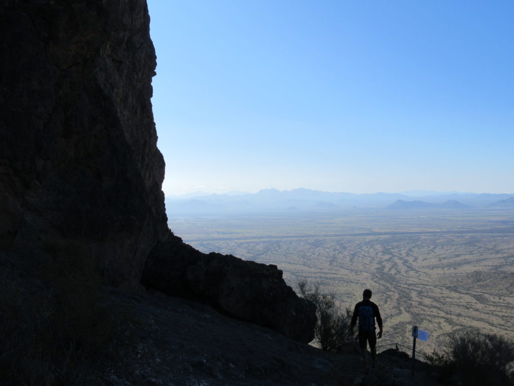 Dark cliff fills half the image with silouette of man walk high above desert valley floor