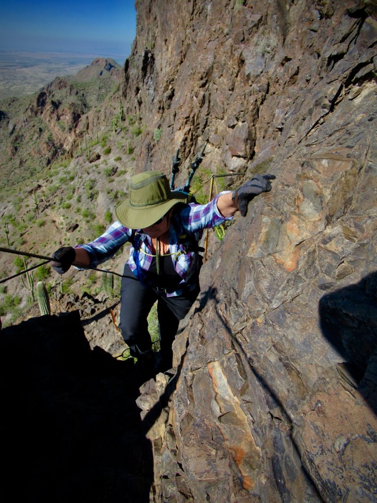 Woman wearing khaki Tilly hat, a plaid shirt, black pants and gloves is holding on to cable with one hand and rock face with the other as she ascends the trail.