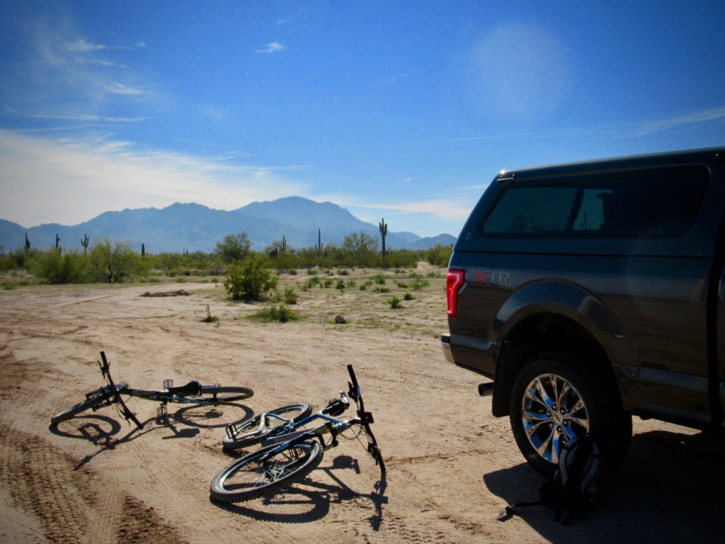 Two mountain bikes lying on sandy road next to grey pickup truck with mountains in distance.