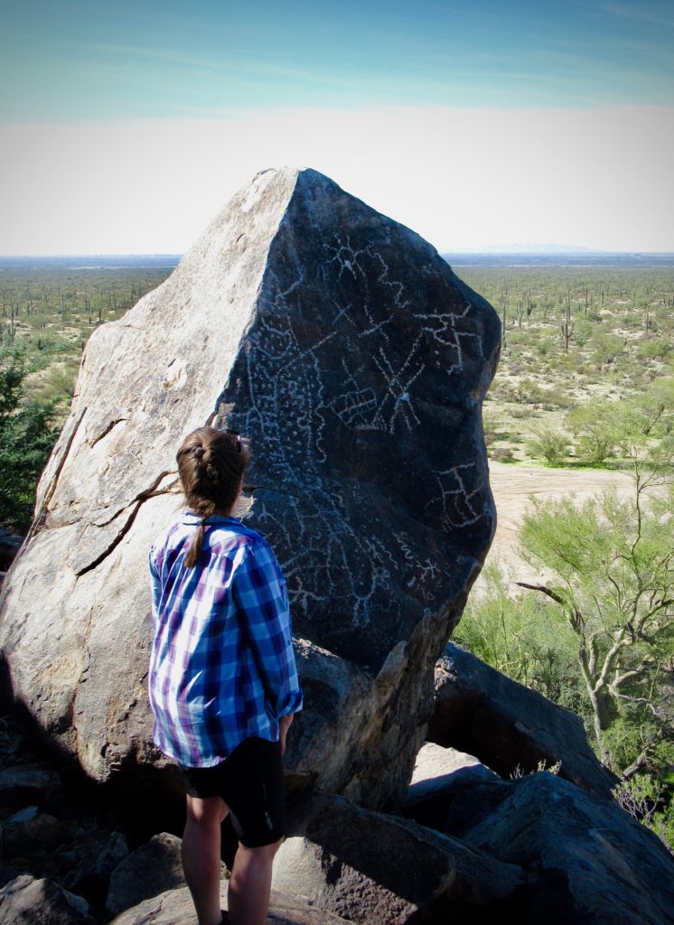 Woman in plaid shirt standing looking at large rock boulder covered in rock art. Desert vegetation in background.