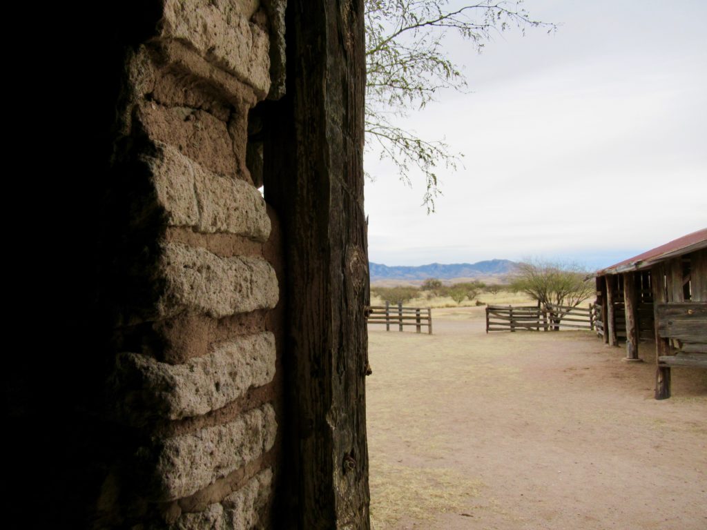 A corner of a weathered building made of adobe bricks and timber blocks view into open ranch years with buildings and fences