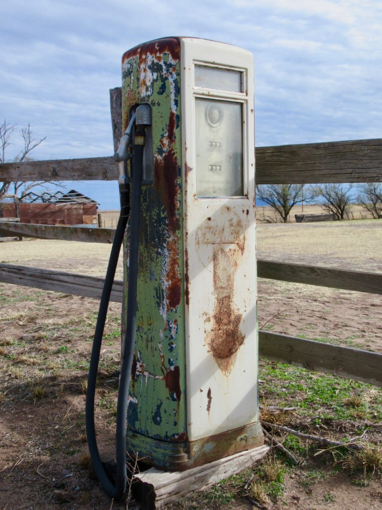 Old, weathered gas pump showing many layers of paint stands in front of a wood fence in a barnyard.

