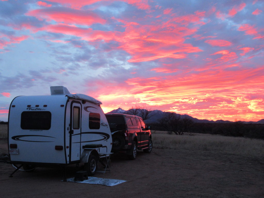 Truck and small travel trailer set up in grassland area with brilliant pinkish-orange sunset glow on scattered clouds overhead.