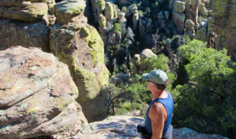 Man with ball cap, blue short and shorts hiking on path above rocky spires in tree-filled canyon in Chiricahua National Monument