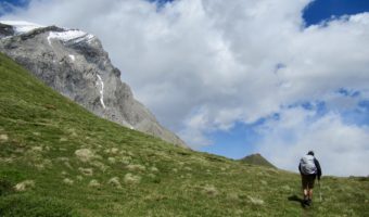 Man in shorts with hiking pole and grey backpack climbing up grassy slope to mountain pass