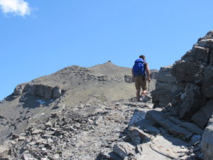 Man with blue pack climbing up rocky ridge with wooden fire lookout in distance