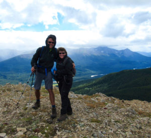 Two people bracing against the wind on a rocky mountain summit with more mountains in distance.