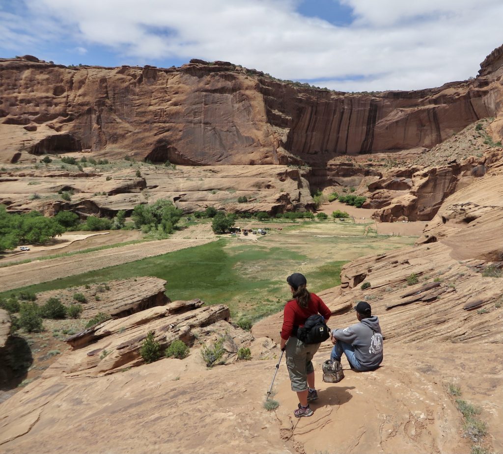 A man sits on a smooth sandstone ledge while a woman in red shirts stands nearby. Both looking out over green fields in canyon bottom.