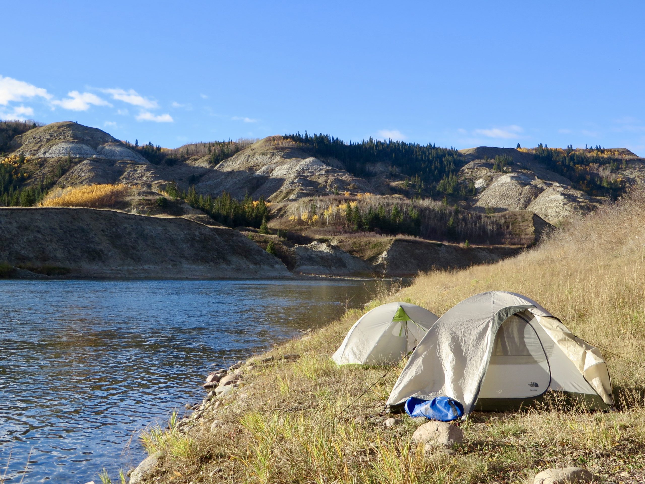 Two tents set up on grassy area beside river with fall colours on trees across the way.