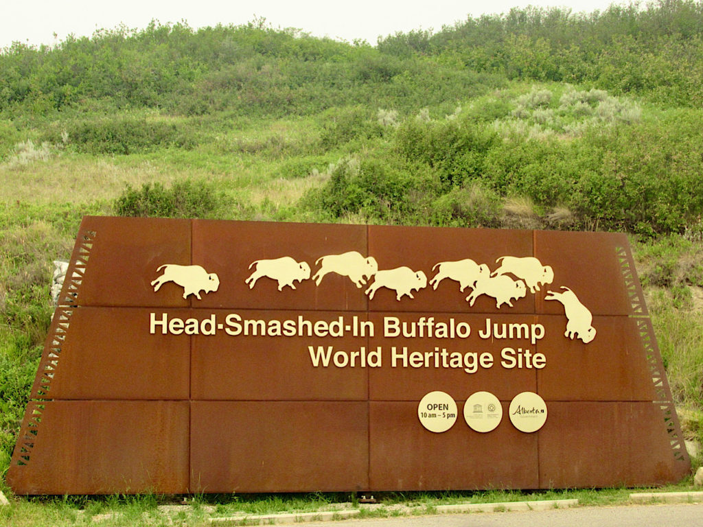 Rusted red metal sign with white bison figures falling from top row with the words "Head-Smashed-In Buffalo Jump World Heritage Site" below.