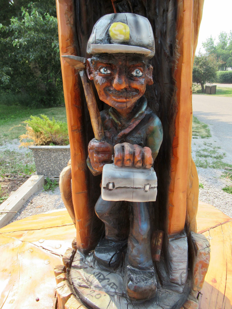Wooden carving of a coal miner wearing hardhat and carrying a pickaxe and lunchbox.