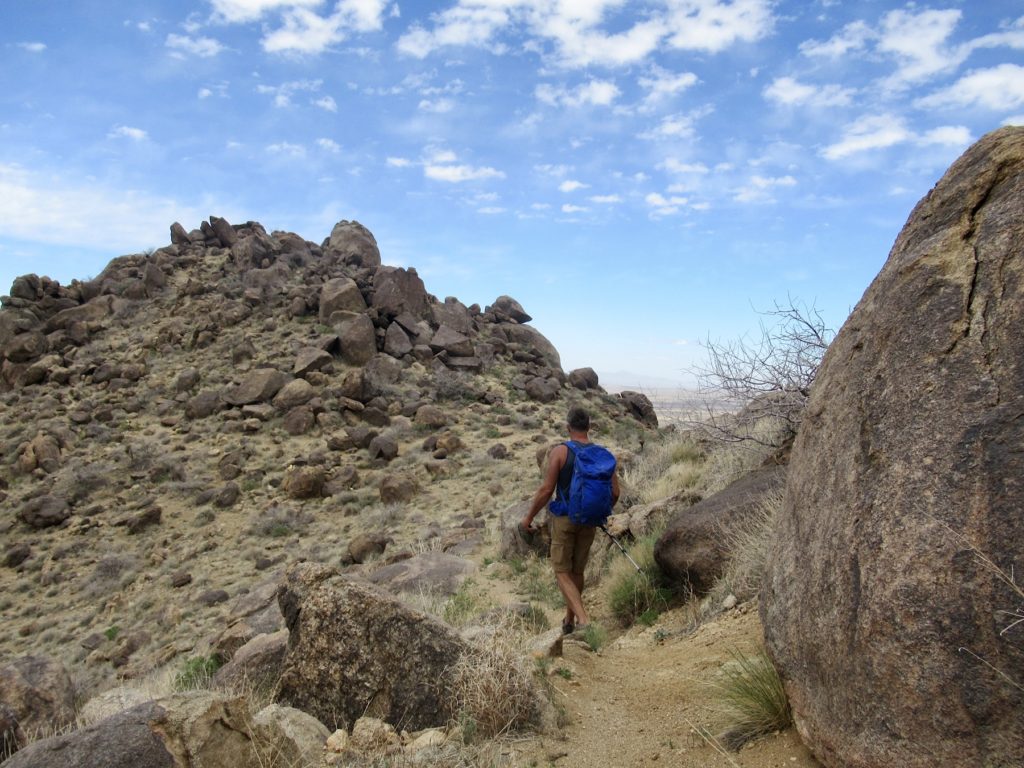Man with blue backpack hiking on a trail surrounded by low rocky mountain peaks.