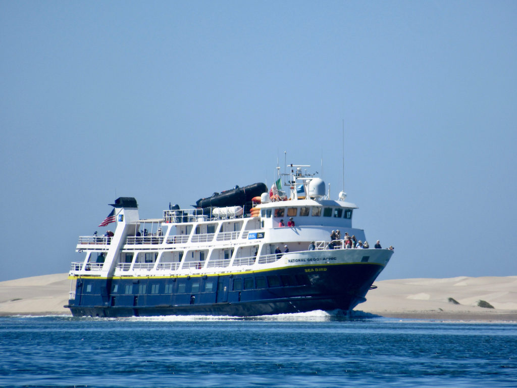 Large cruise ship on blue waters with low sand dunes in background.