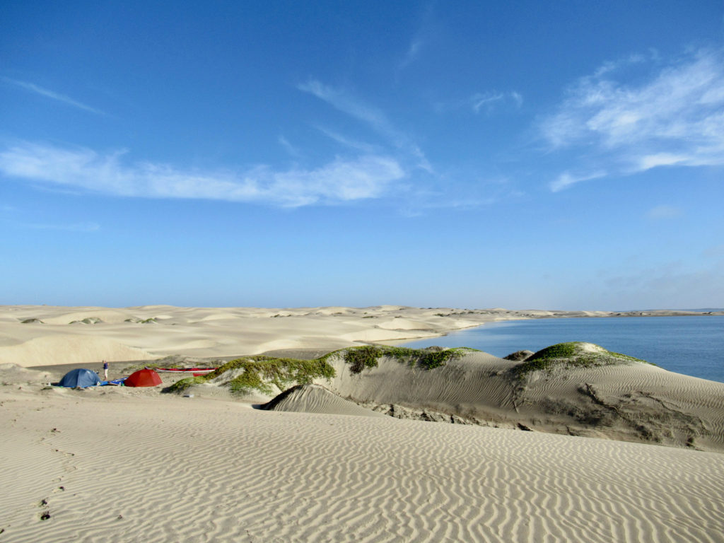 Two small tents (one blue and one red) set up on sand dunes next to water.
