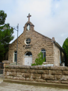 Historic light brown rock church surrounded by green trees with low rock wall in front.