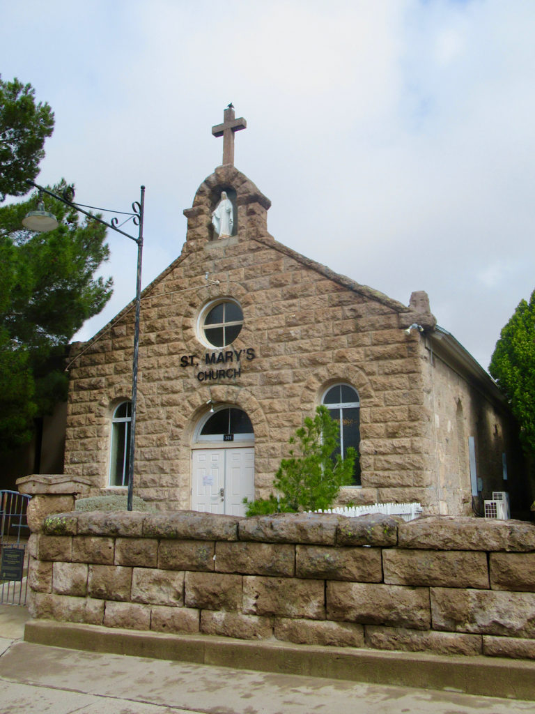 Historic light brown rock church surrounded by green trees with low rock wall in front.